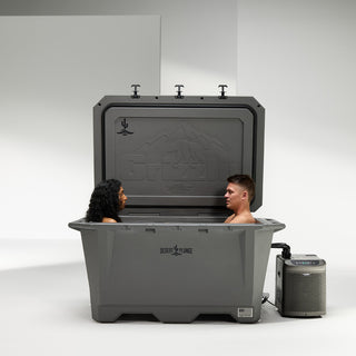 The image shows the Wide cold plunge offering from Desert Plunge in stealth gray. The lid is open on the Grizzly 450 cooler and there is a chiller connected to the iceless ice bath. You can see the Grizzly Coolers logo embossed on the lid and an imprint of the Desert Plunge logo on the top left of the lid. There is also a Desert Plunge logo on the front of the cold plunge tub. Two people are cold plunging at the same time in the Wide cold plunge tub - a man and a woman facing each other. Made in the USA.