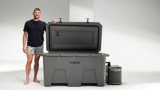 A dark gray cold plunge tub with the lid open with a man standing next to it; he has his left arm on the top of the lid. You can see the Desert Plunge logos on the inside of the lid and on the front center of the cold plunge tub. The man is smiling, wearing patterned swim trunks and a black t-shirt. He has tattoos above the elbow on his right arm.