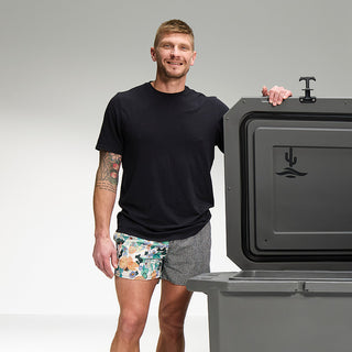 A dark gray cold plunge tub with the lid open with a man standing next to it; he has his left arm on the top of the lid. You can see the Desert Plunge logos on the inside of the lid and on the front center of the cold plunge tub. The man is smiling, wearing patterned swim trunks and a black t-shirt. He has tattoos above the elbow on his right arm.