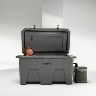 A man using a gray cold plunge tub with his eyes closed, calmly experiencing a deep soak. The cold plunge is sitting indoors in a plain white room. The lid is open as the man is inside. You can see the Desert Plunge logo on the lid and front of the ice bath. The chiller equipment with digital display is hooked up next to the cold plunge.
