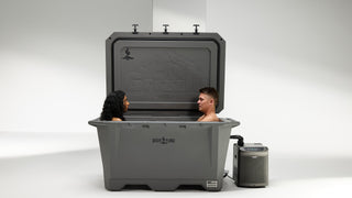 The image features a Desert Plunge Wide 450 Grizzly cold plunge tub with two people cold plunging together. The giant Grizzly 450 cooler is so spacious the couple cold plunging have plenty of room. You can see the Grizzly logo embossed on the lid of the hard sided cooler and imprints of the Desert Plunge logo on the lid and front center of the gray cooler. A chiller is hooked up and operating next to the cold plunge tub.