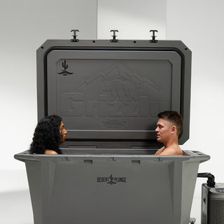 The image features a Desert Plunge Wide 450 Grizzly cold plunge tub with two people cold plunging together. The giant Grizzly 450 cooler is so spacious the couple cold plunging have plenty of room. You can see the Grizzly logo embossed on the lid of the hard sided cooler and imprints of the Desert Plunge logo on the lid and front center of the gray cooler. A chiller is hooked up and operating next to the cold plunge tub.