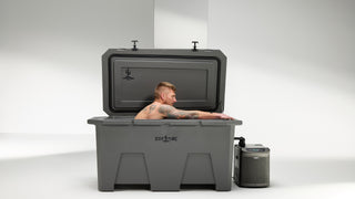 The image shows a man sitting inside a gray cold plunge tub with his arms stretched forward. The lid is open on the cold plunge with a chiller hooked up and operating at the side of the unit. The ice bath is sitting in a white room as to focus on the cold plunge. You can see the Desert Plunge logo on the lid and front of the cold plunge tub which is a Grizzly cooler and is made in the USA.