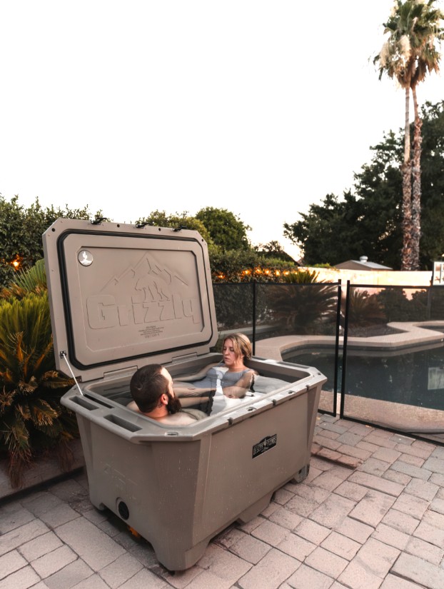 A photo of a man and woman cold plunging together in a Desert Plunge 450 Wide Cold Plunge outside near a pool with palm trees.