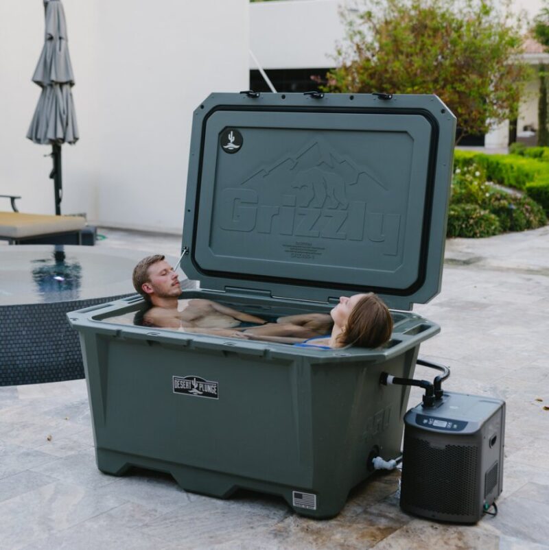 A photo of a Desert Plunge 450 Wide cold plunge outside on a stone patio with the lid open, next to a pool, and two people cold plunging at the same time.